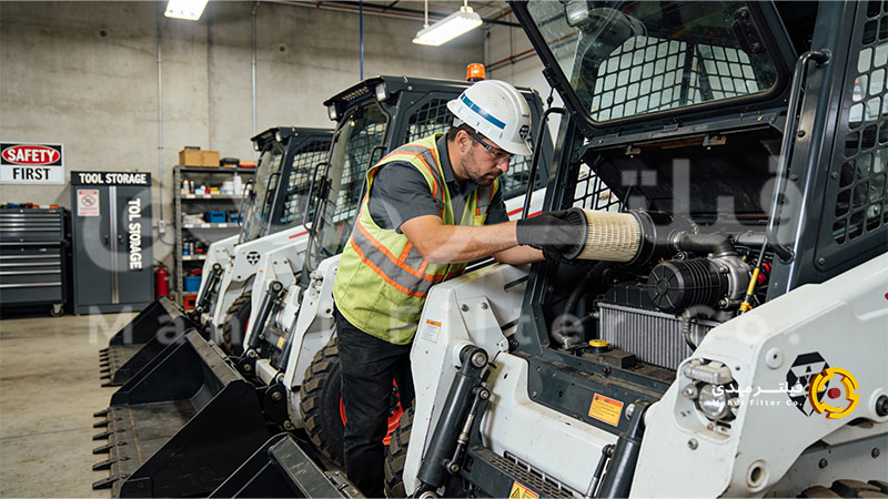 نمای داخلی یک کارگاه یا محوطه صنعتی که در آن چند مینی لودر در کنار هم پارک شدهاند و یک تکنسین در حال چک کردن فیلتر هوای یکی از آنهاست